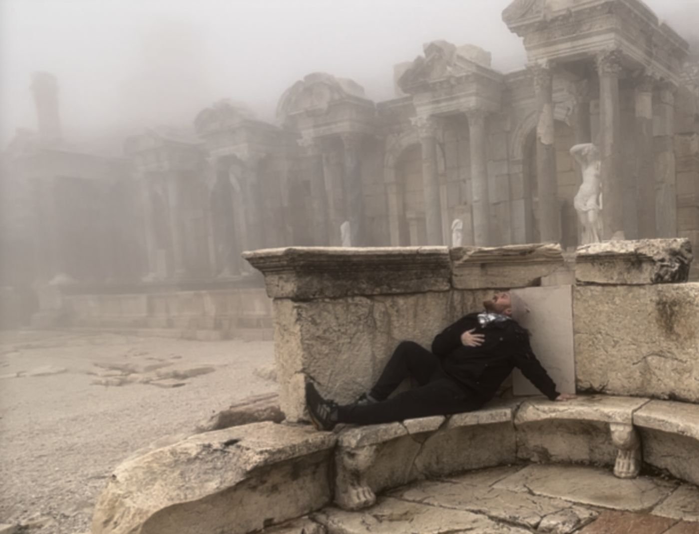 Eric reclining on ancient stone ruins in thick fog, statues and columns behind him