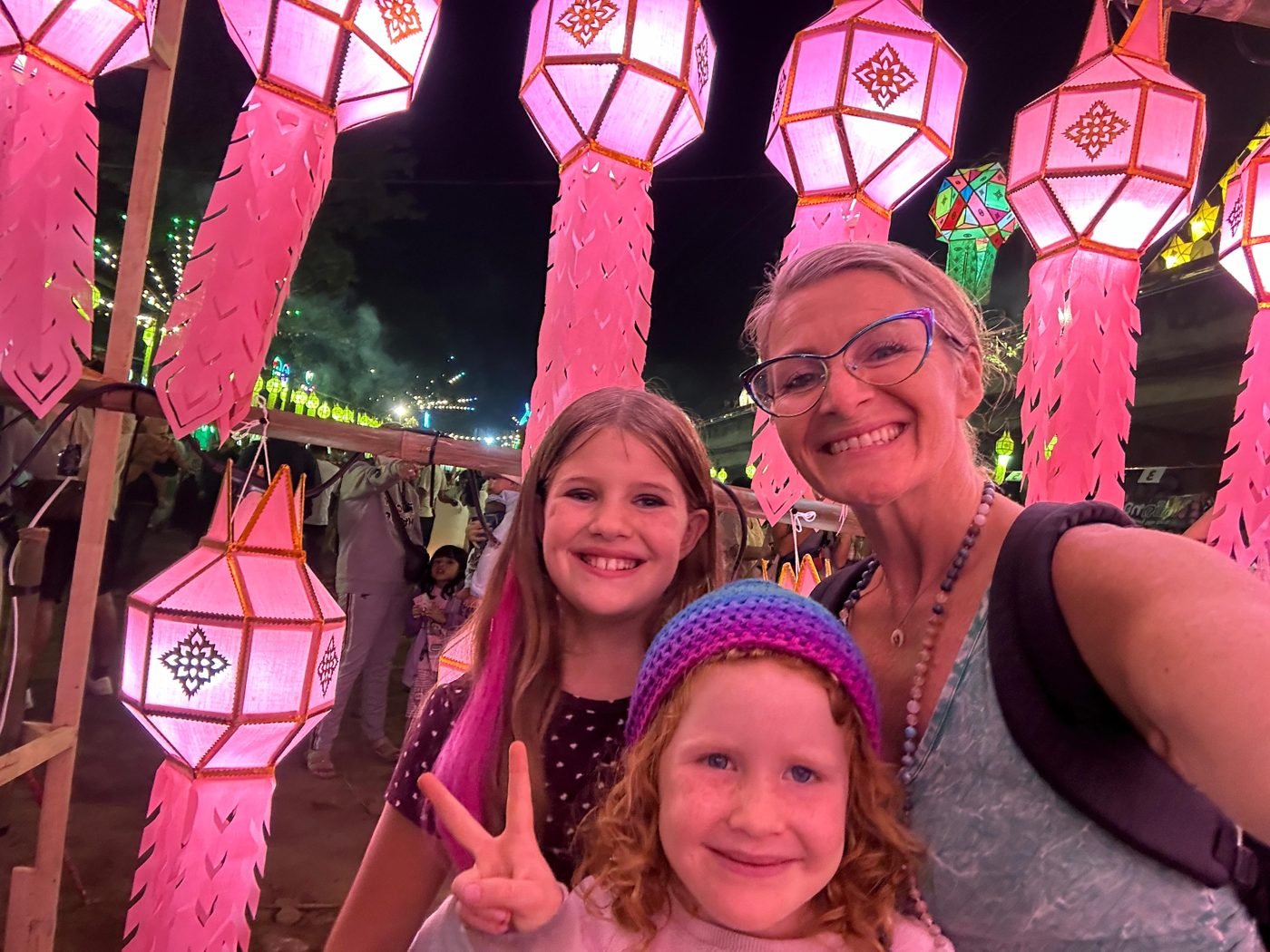 Maya Lila Di Vento and her two daughters smiling under glowing pink lanterns at the Lantern Festival in Pai, Thailand